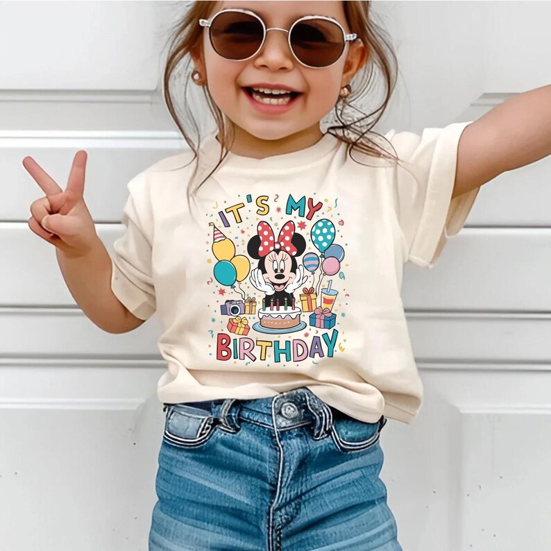 May include: A cream-colored t-shirt with a colorful graphic featuring Minnie Mouse, balloons, a cake, and the text "IT'S MY BIRTHDAY". The shirt is worn by a child wearing blue jeans and sunglasses.