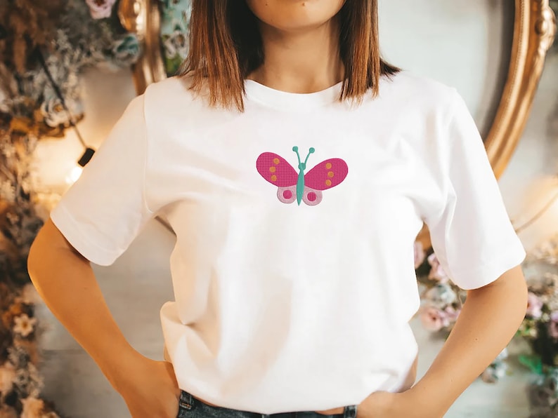 May include: White t-shirt with a pink butterfly embroidered on the front. The butterfly has orange and green accents.