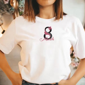 May include: White t-shirt with a black and pink graphic design of the number 8 with a woman's face inside the number and a flower on top. The text "March" is written in pink cursive below the number.