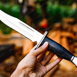 Op de afbeelding: Een groot, gepolijst jachtmes met een glanzend zilveren lemmet en een zwarte handgreep. Het mes heeft een constructie met volle tang en een gebogen lemmet. De handgreep is gemaakt van een donker materiaal. Het mes wordt in een hand gehouden.