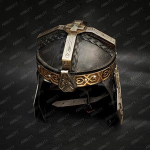 Peut inclure: Casque orné avec une croix sur le dessus, avec des détails dorés et argentés sur une surface sombre et texturée. Le casque présente une bande décorative et une section en cuir tressé. Thème historique ou fantastique.