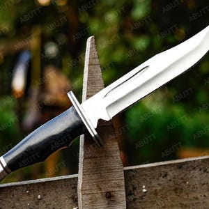 Op de afbeelding: Een groot jachtmes van roestvrij staal met een zwarte handgreep en zilveren accenten. Het lemmet is scherp en schuin, en het mes ligt op een houten hek. Het mes is ongeveer 30 cm lang.