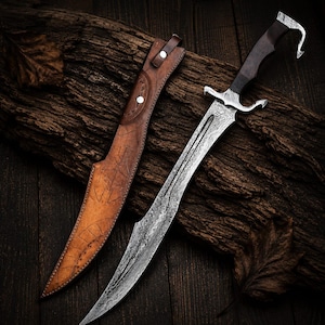 May include: A Damascus steel dagger with a dark wooden handle featuring a curved pommel and a silver-toned crossguard. The blade has a distinct wavy pattern. Beside the dagger is a tan leather sheath with embossed geometric line designs.