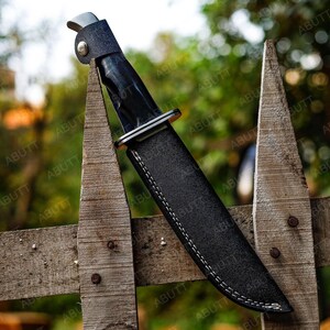 Op de afbeelding: Een zwart jachtmes met een leren schede, rustend op een verweerde houten hek. Het mes heeft een glanzend zwart handvat en een zilveren bolster. De schede is zwart met witte stiksels.