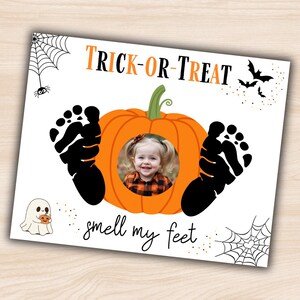May include: Halloween-themed photo frame with a smiling child's picture inside an orange pumpkin. Black footprints flank the pumpkin, with the words "TRICK-OR-TREAT" and "smell my feet" in orange and black. Spiders, webs, bats, and a ghost complete the design.