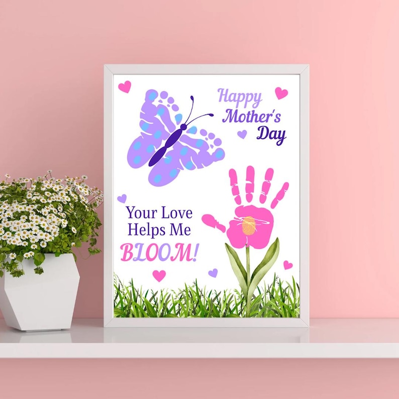 May include: A framed print with a white border features a butterfly and a handprint flower design. The text reads "Happy Mother's Day" and "Your Love Helps Me BLOOM!" in purple and pink. Green grass is at the bottom.