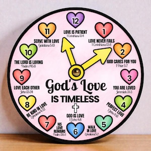 May include: A round clock with a pink background and a black border. The clock face features colorful heart-shaped numbers and phrases like "God's Love is Timeless." The clock hands are yellow, and the text includes Bible verses.