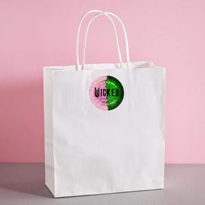 May include: A white paper gift bag with white handles. A circular sticker is affixed to the front, featuring the word "Wicked" in pink and green glitter, with additional text. The background is a solid pink.