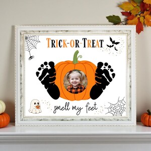 May include: A framed Halloween-themed print with a white border. The print features a pumpkin with a child's photo in the center, black footprints, spiderwebs, bats, and the text "TRICK-OR-TREAT" and "smell my feet".
