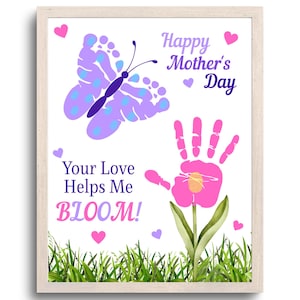 May include: A framed print with a white background features a butterfly and a flower made from handprints. The text reads "Happy Mother's Day" and "Your Love Helps Me BLOOM!" in purple and pink. Green grass is at the bottom.