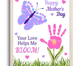 May include: A framed print with a white background features a butterfly and a flower made from handprints. The text reads "Happy Mother's Day" and "Your Love Helps Me BLOOM!" in purple and pink. Green grass is at the bottom.