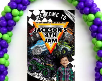 May include: A party sign with the text "WELCOME TO THE MONSTER TRUCK BASH! JACKSON'S 4TH JAM" featuring colorful monster truck illustrations. The sign is on a wooden easel, with a balloon arch in green and purple.