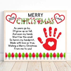 May include: A framed Christmas print with the text "Merry Christmas" and a red handprint with a heart. The print includes the name "Layla" and the year "2025". A string of red and green lights decorates the bottom.