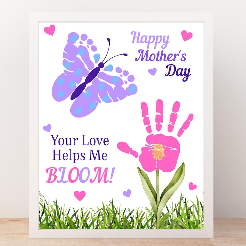 May include: A framed print with a white border features a butterfly made of footprints and a handprint flower. Text reads "Happy Mother's Day" and "Your Love Helps Me BLOOM!" in purple and pink. Green grass and pink hearts decorate the bottom.
