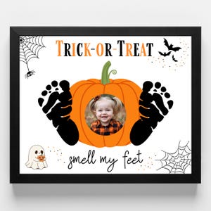 May include: Black framed Halloween-themed print with a pumpkin design. The pumpkin has a photo of a child in the center, with black footprints on either side. The words "Trick or Treat" and "smell my feet" are written on the print, along with spiderwebs, bats, and a ghost.