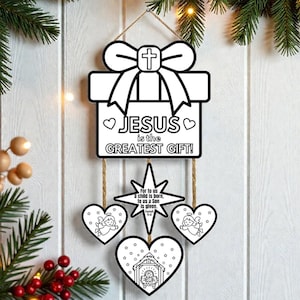 May include: A white wooden Christmas decoration in the shape of a gift box with a cross and the text "JESUS is the GREATEST GIFT!". Below are star and heart-shaped ornaments with religious imagery. The decoration hangs from a jute string.