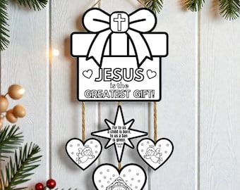 May include: A white wooden Christmas decoration in the shape of a gift box with a cross and the text "JESUS is the GREATEST GIFT!". Below are star and heart-shaped ornaments with religious imagery. The decoration hangs from a jute string.