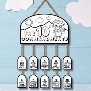 May include: A black and white wooden sign displaying "The 10 Commandments." The sign features a cartoon shepherd illustration and lists the ten commandments below. It is suspended by a brown rope, suitable for hanging. The sign is made of wood.