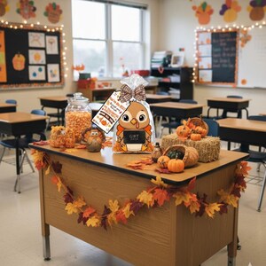 May include: A classroom desk is decorated for fall with a gift bag featuring a cartoon turkey holding a chalkboard that says "GIFT". The desk also has pumpkins, hay bales, and a garland of autumn leaves. A jar of candy corn is visible.