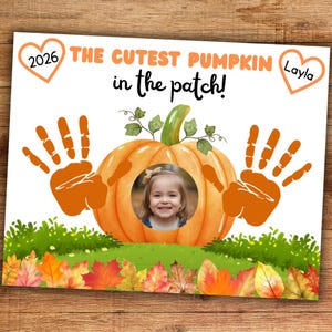 May include: A rectangular sign featuring the text THE CUTEST PUMPKIN in the patch! with a central orange pumpkin illustration containing a circular photo of a child. Two orange handprints flank the pumpkin, with hearts labeled 2026 and Layla.
