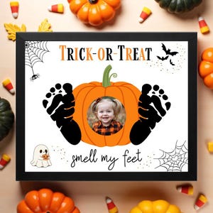 May include: A framed Halloween-themed print with the words "Trick or Treat" in orange and black. A pumpkin shape contains a baby's photo, with black footprints on either side. The phrase "smell my feet" is written below.