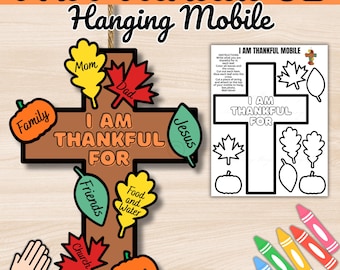 Puede incluir: Móvil colgante en forma de cruz de madera marrón con el texto «I AM THANKFUL FOR». Presenta recortes coloridos de hojas y calabazas con palabras como «Familia», «Amigos» y «Hogar». Incluye una página para colorear y crayones.