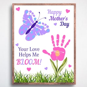 May include: A framed print with a white background features a butterfly made of footprints and a handprint flower. Text reads "Happy Mother's Day" and "Your Love Helps Me BLOOM!" in purple and pink. Green grass and pink hearts decorate the bottom.