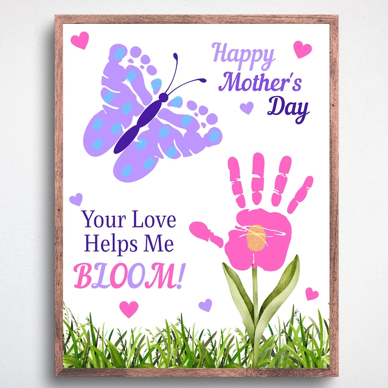 May include: A framed print with a white background features a butterfly made of footprints and a handprint flower. Text reads "Happy Mother's Day" and "Your Love Helps Me BLOOM!" in purple and pink. Green grass and pink hearts decorate the bottom.
