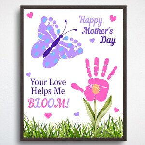 May include: A framed print with a white background features a butterfly made of footprints and a handprint flower. Text reads "Happy Mother's Day" and "Your Love Helps Me BLOOM!" with pink and purple hearts and green grass at the bottom.