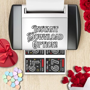 May include: A printer printing Valentine's Day cards with the text "Instant Download Option!". The cards feature the numbers "67" and phrases like "You're the 67 to my..." and "You + Me = 67". A heart-shaped box, candy hearts, and roses are also visible.