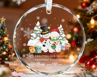 May include: A clear, faceted glass Christmas ornament with a white ribbon. The ornament features two snowmen, Christmas trees, and gift boxes. Text reads "Celebrating Our Second Christmas Together" with names and the year 2025.