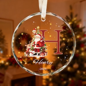 May include: A circular glass Christmas ornament featuring a white ribbon hanger. The design includes a large red letter H, a graphic of Santa Claus with a reindeer and snowman, the name Henry, and the year 2025, surrounded by gold stars.