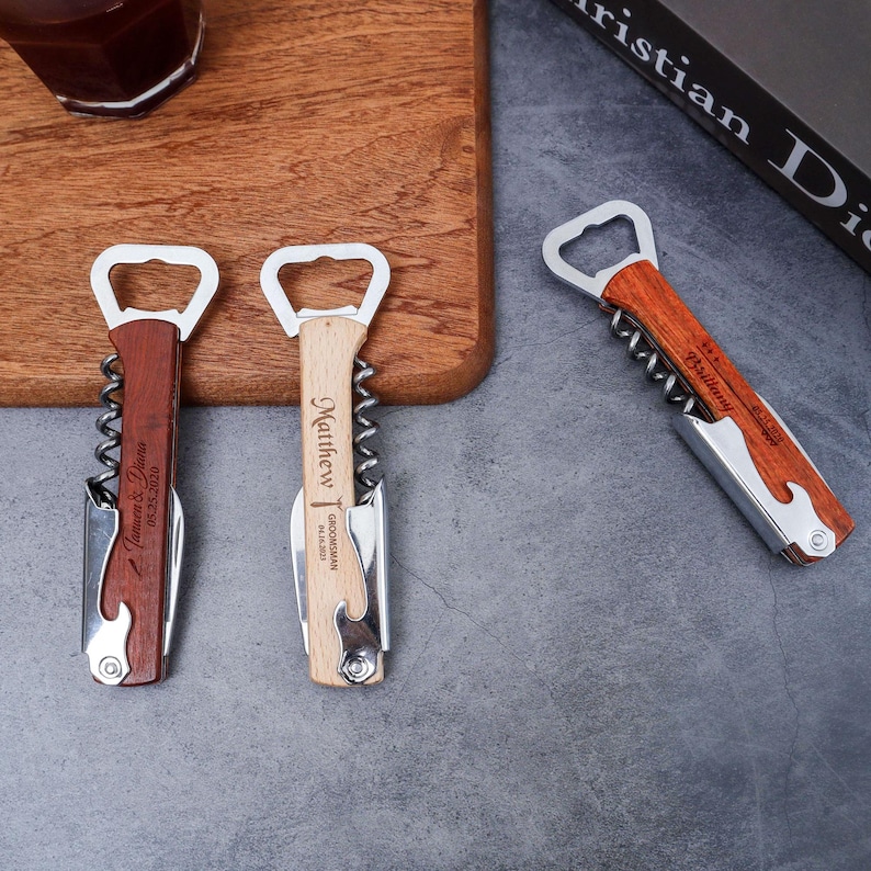 May include: Three personalized wooden bottle openers with corkscrews and bottle cap openers. Two openers are on a wooden cutting board, and one is on a gray surface. Each opener has a different name and date engraved on the wooden handle.