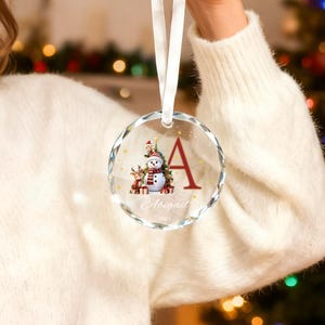 May include: A round, faceted glass Christmas ornament hanging from a white ribbon. The ornament features a printed graphic of a snowman, a reindeer, and a Christmas tree, alongside a large red letter A, the name Abigail, and 2025.