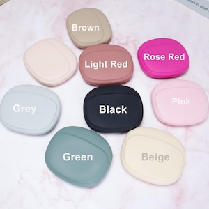 Op de afbeelding: Een verzameling van negen beschermhoezen voor oordopjes in verschillende kleuren, waaronder bruin, lichtrood, rozerood, grijs, zwart, roze, groen en beige. Elke hoes heeft een afgeronde vorm met de kleurnaam erop.