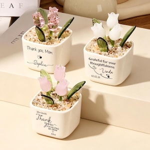 Può includere: Tre vasi quadrati in ceramica bianca riempiti con piccole pietre e tulipani in vetro. Uno riporta la scritta Thank you, Mom. Sophia, un altro Grateful for your thoughtfulness Linda 02.02.26, e il terzo Mrs Smith Thank you FOR HELPING ME GROW Alice.