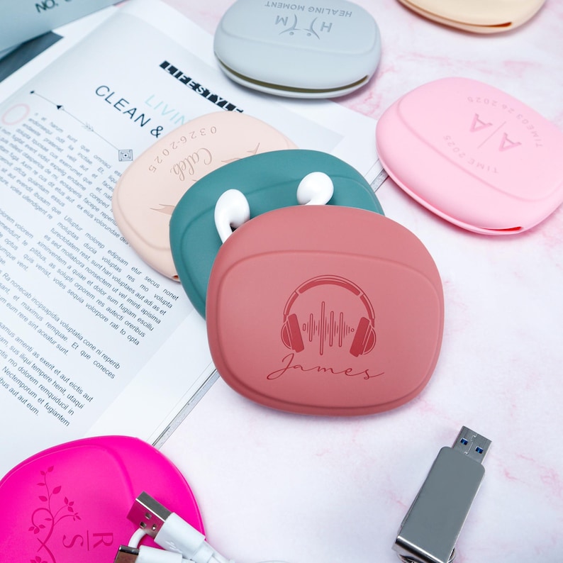 Op de afbeelding: Een verzameling kleurrijke oortelefoonhoesjes in roze, teal, perzik en grijs. E&eacute;n hoesje is gegraveerd met een koptelefoonontwerp en de naam "James". Andere hoesjes hebben de tekst "Healing Moment", "Caleb" en "Time 2025".