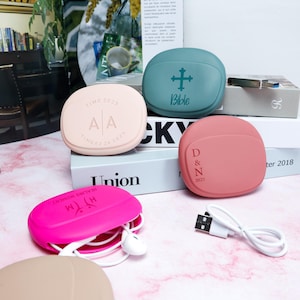May include: A selection of colourful, rounded earbud cases. The cases are in shades of pink, teal, and peach, with text and graphics on them. One case is open, revealing white earbuds. A white USB cable is also present.