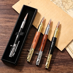 Könnte beinhalten: Eine Sammlung von kugelförmigen Stiften, einer in einer schwarzen Box und drei lose. Die Stifte haben metallische Oberflächen mit goldenen, silbernen und schwarzen Details. Die Holzstifte sind mit Namen graviert. Auf einer Holzoberfläche.