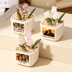 Op de afbeelding: Drie vierkante witte keramische plantenbakken gevuld met kleine steentjes en glazen tulpen. Elke pot is voorzien van een aangepaste foto en tekst: 'love you forever', 'Happy birthday' en 'love you'. De tulpen zijn roze of wit met groene bladeren.