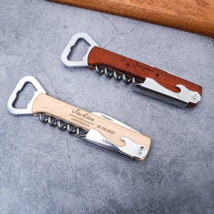 May include: Two personalized bottle openers with corkscrews. One has a light wood handle with the name "Jackson" and the date "05.20.2023". The other has a dark wood handle with the name "Sebastian" and the date "05.20.2024".