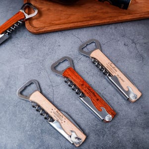May include: A collection of personalized wooden wine bottle openers with stainless steel components. Each opener features a corkscrew, bottle opener, and a small knife. The openers are engraved with names and dates, perfect for groomsmen gifts.