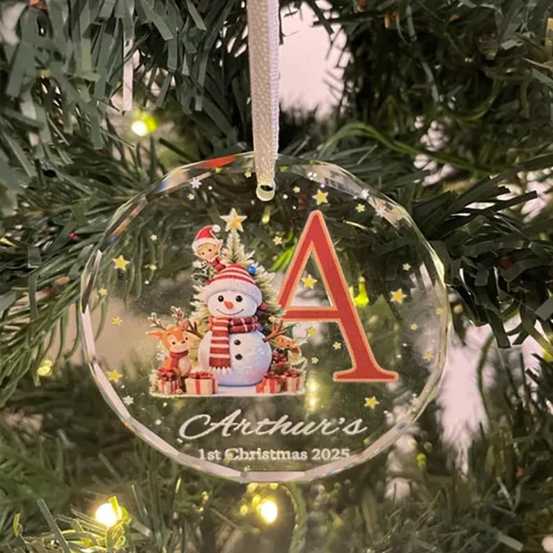 May include: A clear, circular glass Christmas ornament featuring a large red letter A, a snowman with a red and white striped scarf, a small reindeer, and a tiny elf. The text reads Arthur's 1st Christmas 2025, accented with yellow stars.