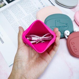 Op de afbeelding: Een felroze siliconen etui bevat een witte oplaadkabel. Op de achtergrond zijn verschillende andere siliconen hoesjes in verschillende kleuren te zien, waaronder turquoise en roze. De turquoise hoes heeft een Texas-omtrek en tekst. De hoesjes lijken voor oortelefoons te zijn.
