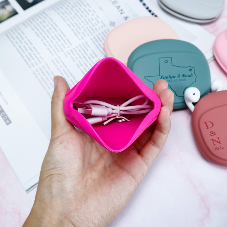 Op de afbeelding: Een felroze siliconen etui bevat een witte oplaadkabel. Op de achtergrond zijn verschillende andere siliconen hoesjes in verschillende kleuren te zien, waaronder turquoise en roze. De turquoise hoes heeft een Texas-omtrek en tekst. De hoesjes lijken voor oortelefoons te zijn.