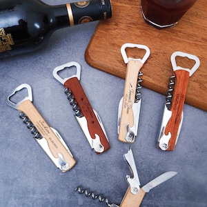 Pode incluir: Uma coleção de abridores de garrafas de vinho de madeira personalizados com saca-rolhas e abridores de garrafas. Cada abridor tem uma pega de madeira clara com um detalhe em madeira escura e texto gravado. Uma garrafa de vinho e um copo de vinho tinto também estão na imagem.