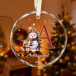 May include: A circular glass Christmas ornament with a faceted edge and a white ribbon hanger. It features a snowman, a reindeer, a Christmas tree, the letter A, the name Abigail, and the year 2025 printed on the surface with gold stars.