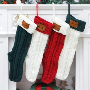 May include: Four cable-knit Christmas stockings in green, red, and white hang from a white mantelpiece. Each stocking has a personalized leather tag with a name or initials. Festive holiday decor includes a Santa figure and greenery.