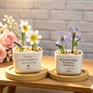 Peut inclure: Deux petits pots carrés en céramique blanche avec des sculptures de fleurs en verre. L'un présente des marguerites blanches, l'autre des iris violets. Chaque pot est posé sur un dessous de verre en bois et porte un message imprimé.