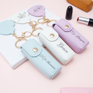 Puede incluir: Llaveros y estuches de pintalabios personalizados en colores pastel. Los llaveros tienen forma de corazón con diseños florales. Los estuches de pintalabios son azul claro, blanco y lavanda, cada uno con un nombre y un detalle floral.